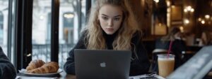 Woman in dark sweater focused on laptop, surrounded by coffee and croissants