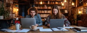 Two college students working on laptops at a rustic library table
