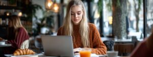 tudent in a sweater working on her laptop at a café table with juice and a croissant.