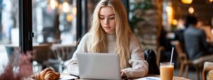 Blonde female student focused on her laptop in a cozy café with coffee and a croissant.