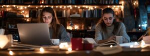 Two students studying in a cozy library with laptops and books around.