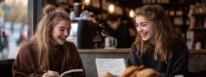 Two college students laughing over study notes and pastries in a warm café.