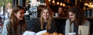 Three college students laughing and talking in a café setting with coffee and notebooks.