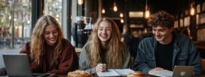 Group of students smiling in a café with laptops, notebooks, and pastries on the table.