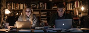 Two college students studying on laptops at a desk surrounded by books and notes in a dimly lit study room.