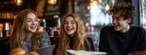 Three college students smiling and laughing together in a café after studying with a laptop.